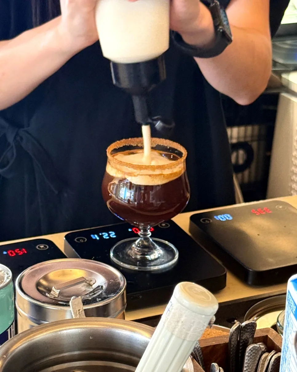 縁にトッピングをまぶしたグラスへクリームを注ぐイオイコーヒー