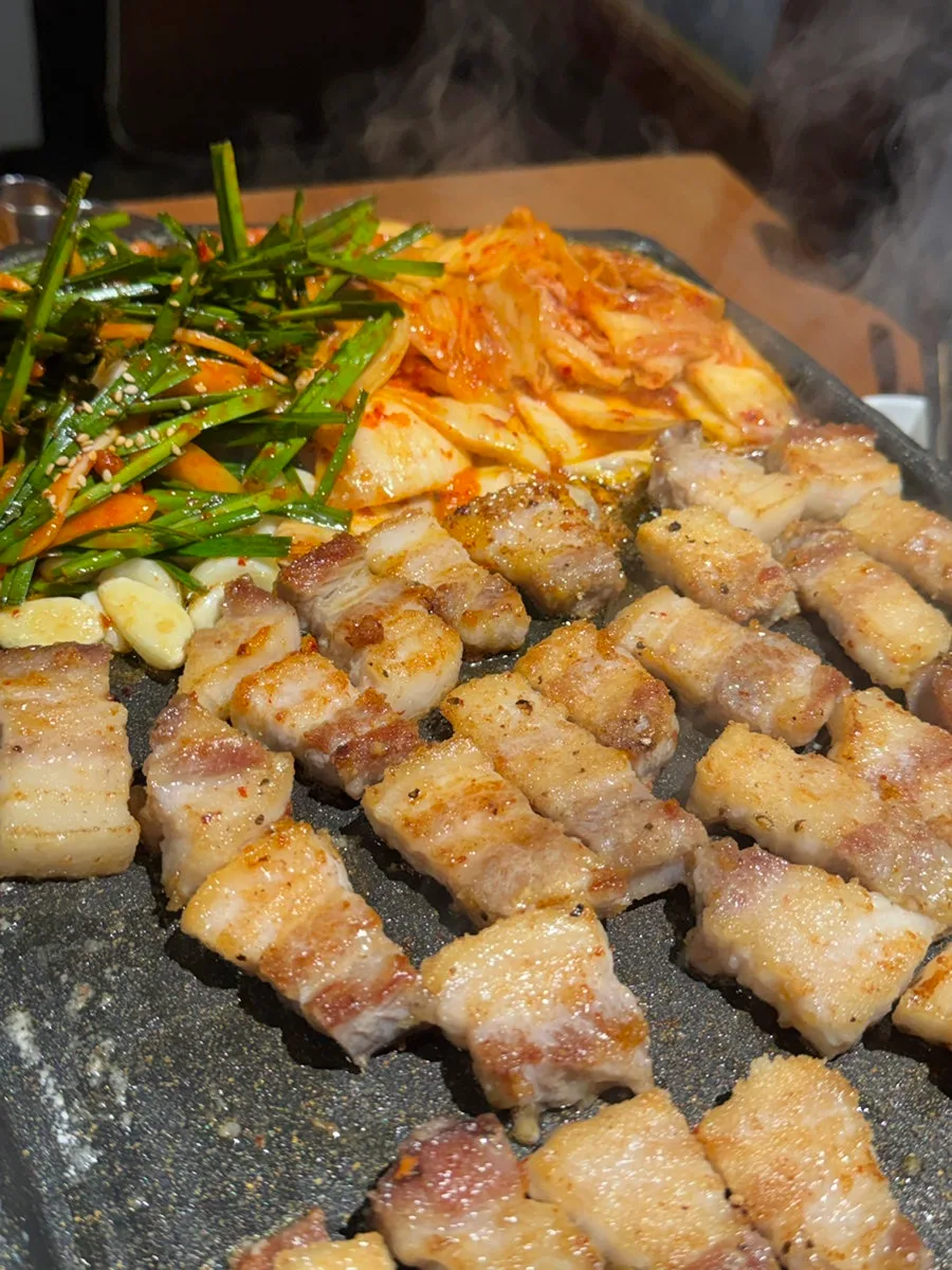 マッキムチ生サムギョプサル 延南店 — ニラの和え物とキムチの横で焼き上がるサムギョプサル