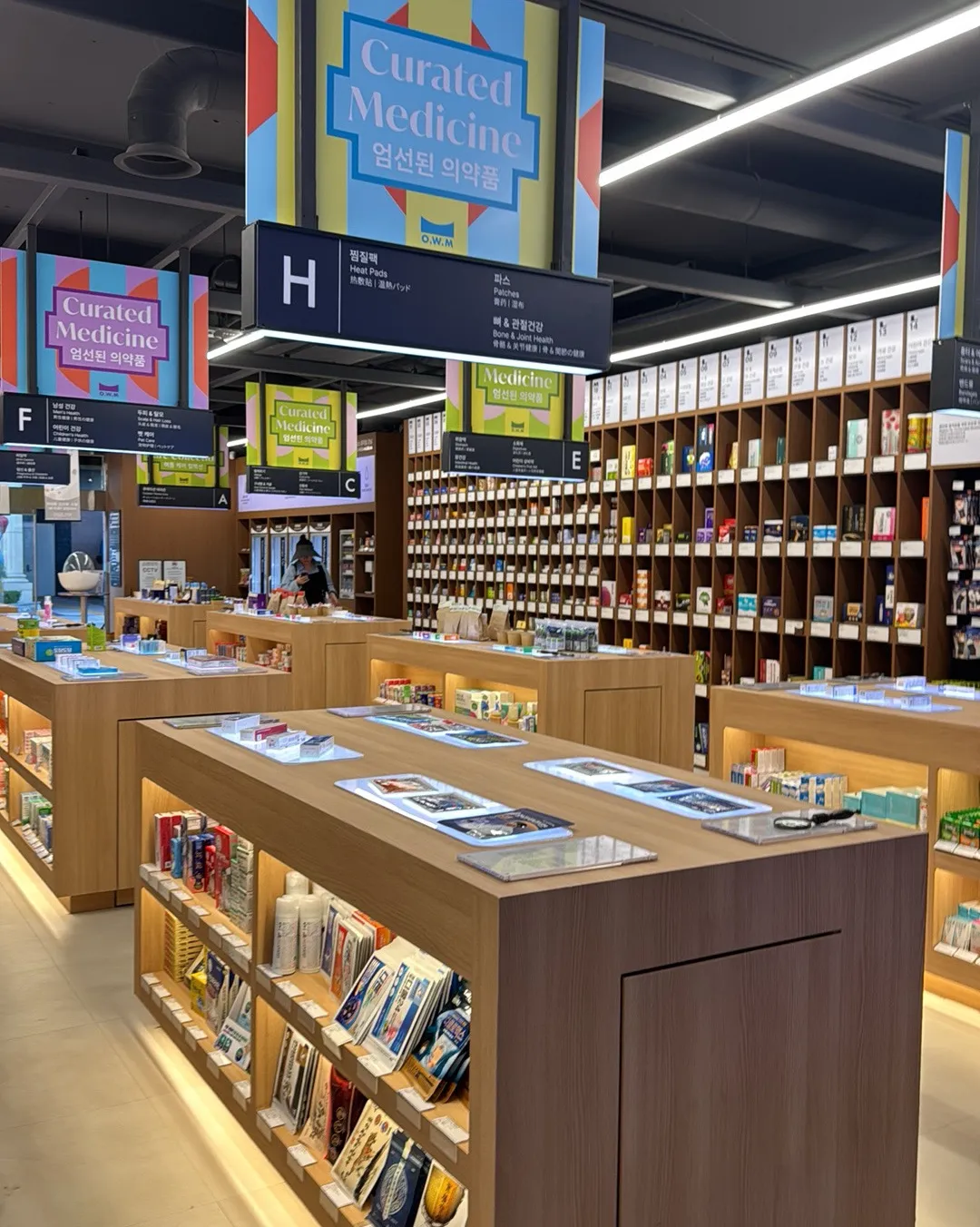 オプティマウェルネスミュージアム薬局 江南店 — 「Curated Medicine」の案内と木の格子棚に並ぶ薬