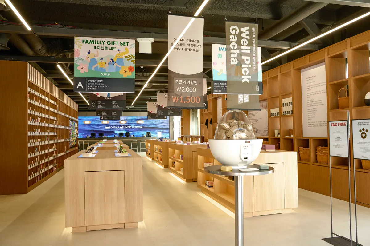 オプティマウェルネスミュージアム薬局 江南店 — ガチャマシンと木の陳列台が並ぶ店内